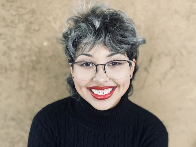 Person with black turtle neck, short dark brown hair with blonde streaks, red lipstick and glasses. 