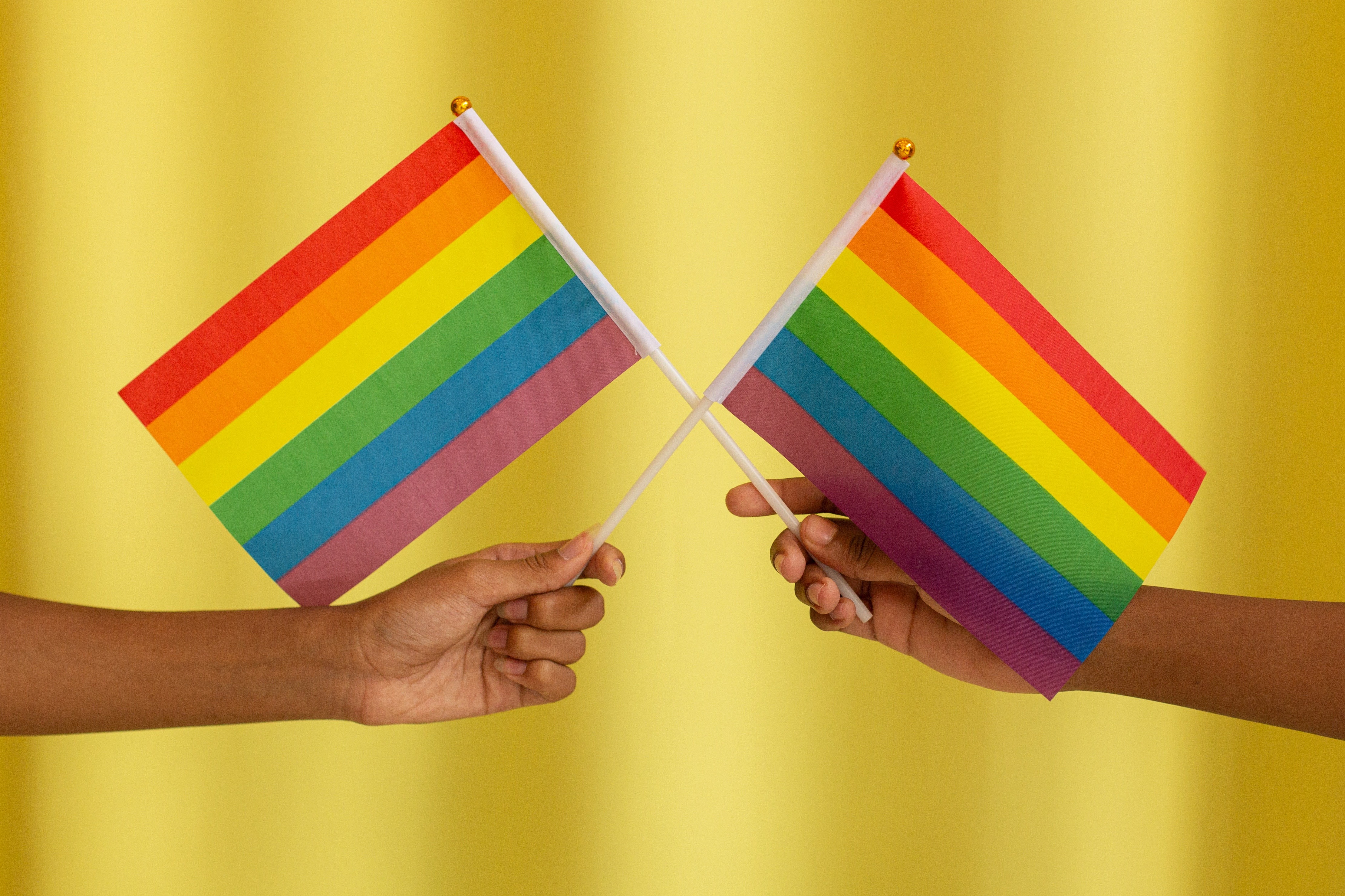 Two hands holding two rainbow flags against a yellow background